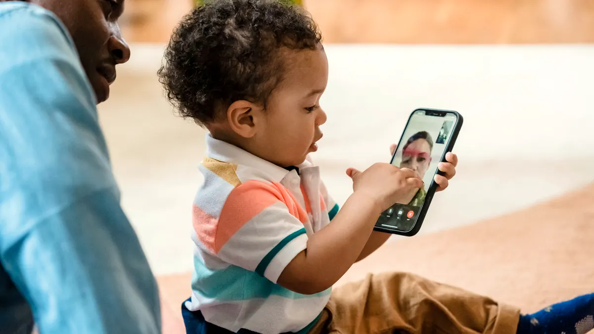 Toddler on a video call with a family member on a smartphone while an adult looks over his shoulder