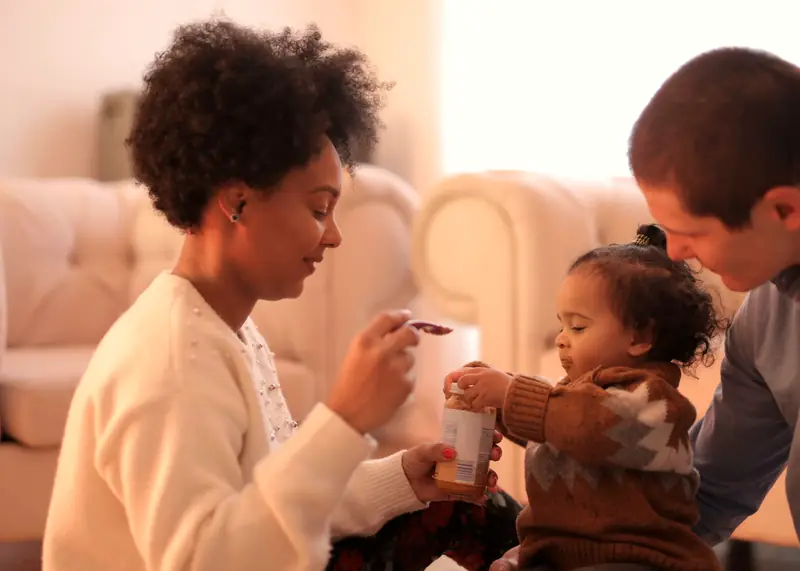 Mom offering a spoonful of food to her toddler, who holds a small jar, while dad looks on in the living room