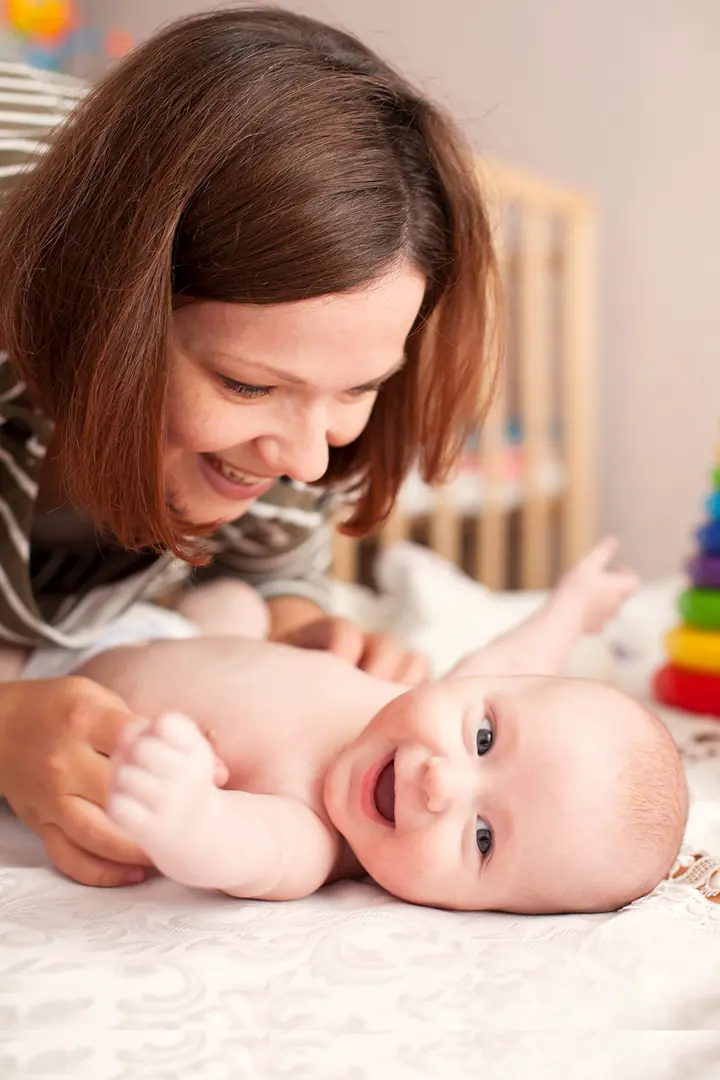 A smiling woman leans over a laughing baby lying on a soft white blanket in a bright, warm nursery