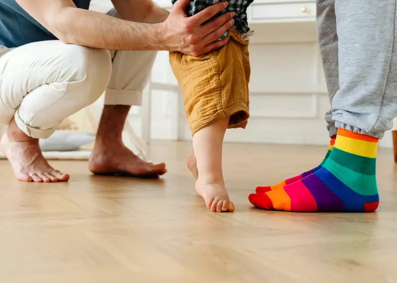 Toddler taking first steps on a herringbone wood floor while an adult steadies him and another stands close by