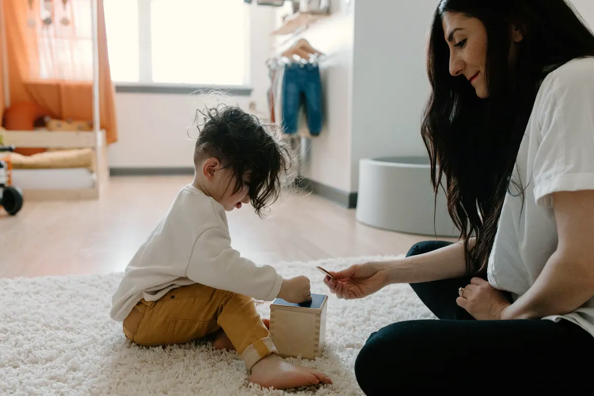 Woman on a soft white rug holding up a small card for a toddler who is playing with a wooden toy