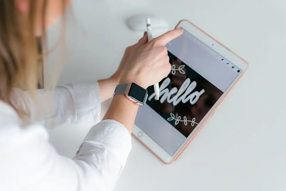 Hands using a stylus on a tablet at a bright white desk, with the word “hello” hand-lettered on the screen