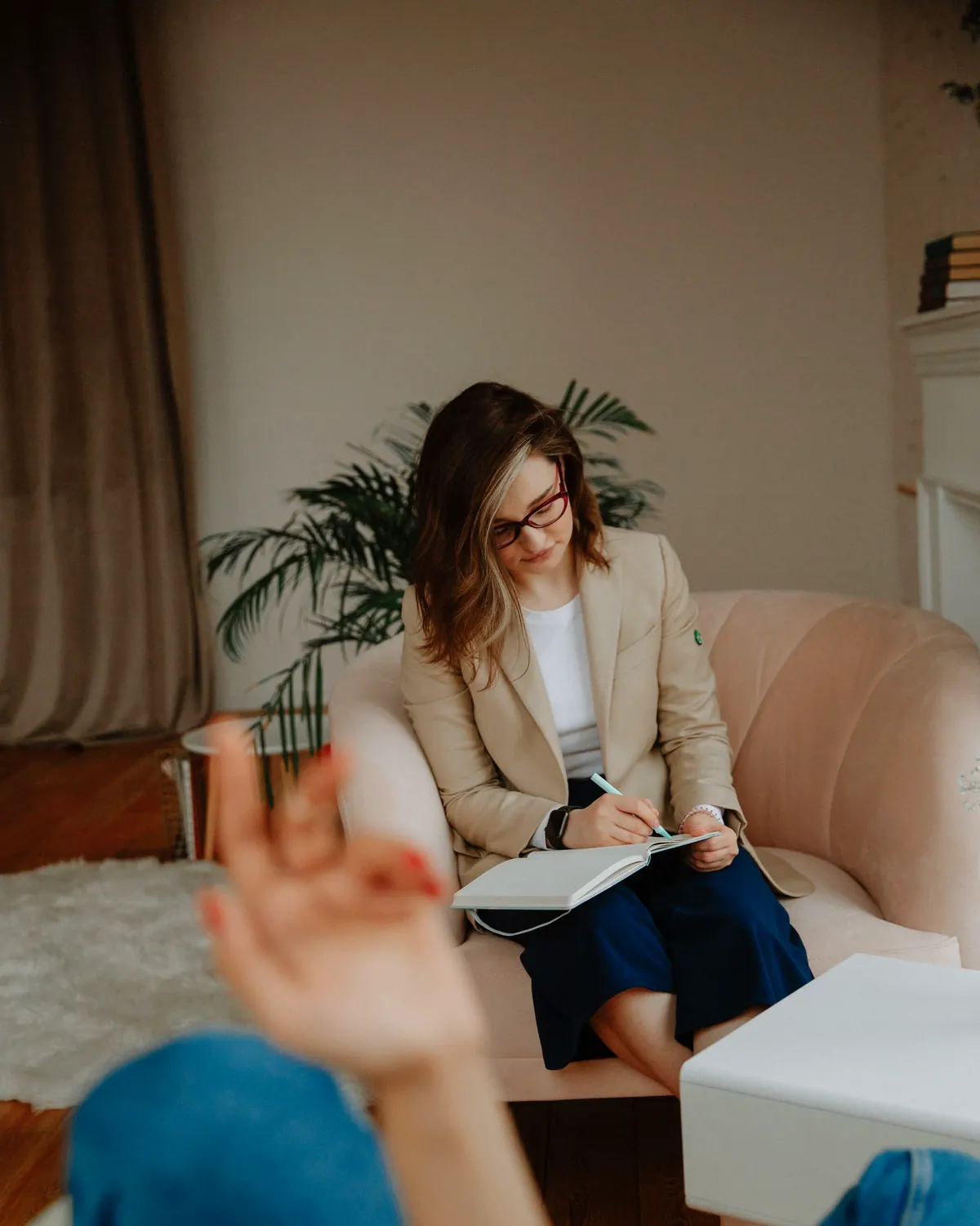 Therapist in a tan blazer taking notes in a notebook while seated in a soft pink armchair in a bright living room