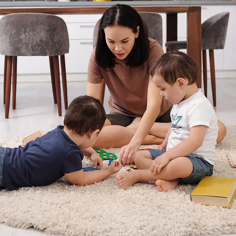 A woman sits on a round rug playing with two toddler boys and wooden puzzles in a bright living room