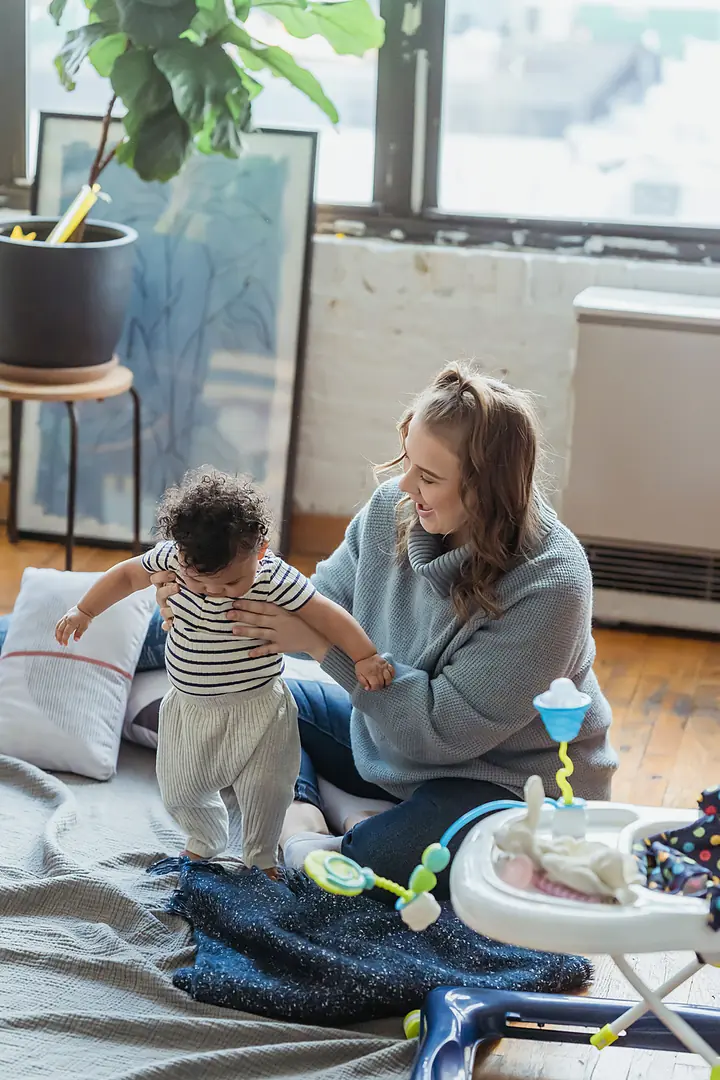 A smiling woman in a light-blue sweater sits on a wood floor and steadies a baby who is standing on a blanket in a bright living room