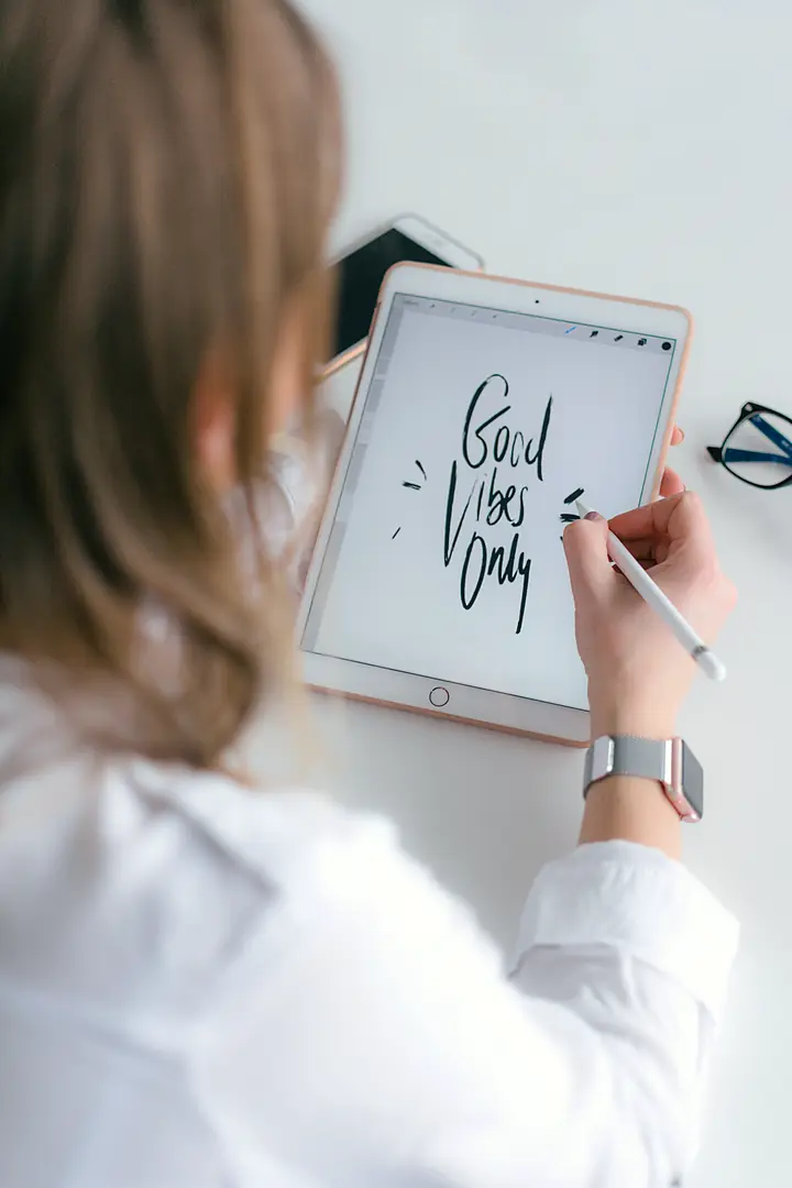 Hands writing with a stylus on a tablet at a bright white desk, with “Good Vibes Only” hand-lettered on the screen